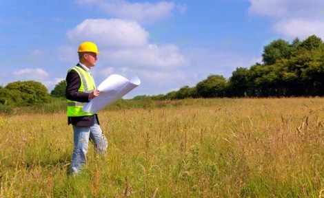 An architect with a yellow hard hat on and high viz jacket is stood in a tree lined field looking at building plans that he is holding in his hand.