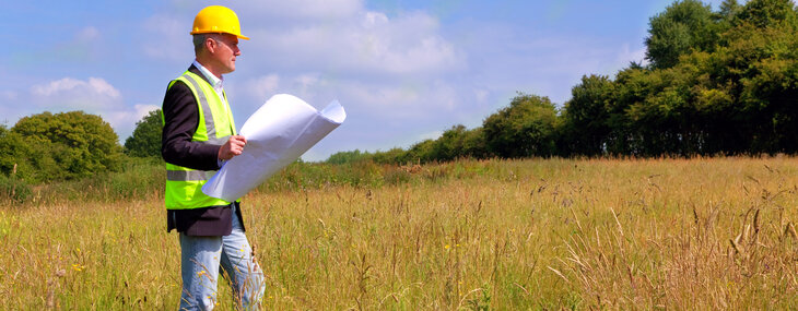 An architect with a yellow hard hat on and high viz jacket is stood in a tree lined field looking at building plans that he is holding in his hand.