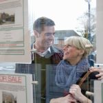 A heterosexual couple are pictured in a warm embrace and smiling as they look into an estate agent window.