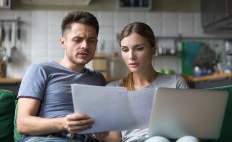 A heterosexual couple are pictured in their kitchen looking at bills to pay while looking at a laptop.