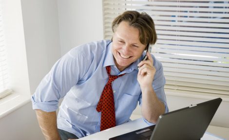 A man is sat at his desk with his sleeved rolled up in a blue shirt and a loose red tie around his neck laughing on the telephone.