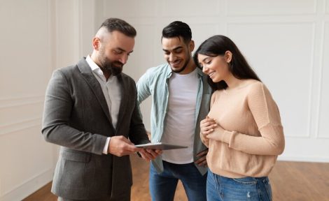 A hybrid estate agents is using a tablet, showing a young hetrosexual couple the floor plans of their potential new home that they are stood in.