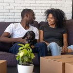 An African American heterosexual couple are pictured with their dog they bought during lockdown sat on a sofa with boxes they have packed to move surrounding them.